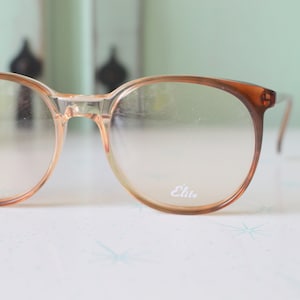 May include: A pair of brown plastic framed glasses with clear lenses. The glasses have the word "Elie" printed on the right lens.