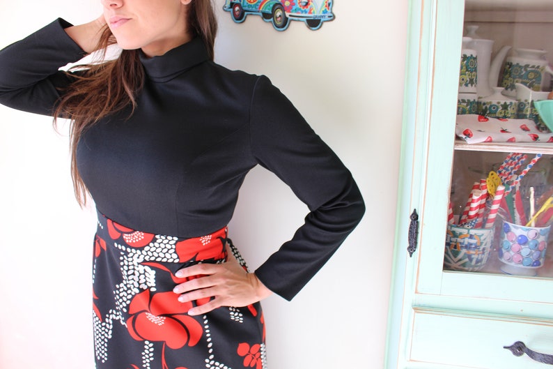 Peut inclure: Haut noir &agrave; manches longues avec un col montant, associ&eacute; &agrave; une jupe avec un fond noir et un motif floral rouge et blanc audacieux. L'ensemble est de style vintage.