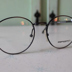 May include: A pair of eyeglasses with round, black frames and clear lenses. The glasses have a thin metal frame and the brand name "GIRBRO" is printed on one of the lenses. The glasses are resting on a white surface.