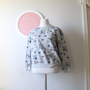 May include: A gray sweatshirt with a floral pattern in blue and purple. The sweatshirt is displayed on a mannequin. The text "Retro & Me Vintage" is visible on a pink and white circular sign in the background.