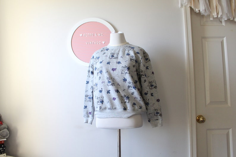May include: A gray sweatshirt with a floral pattern in blue and purple. The sweatshirt is displayed on a mannequin. The text "Retro & Me Vintage" is visible on a pink and white circular sign in the background.