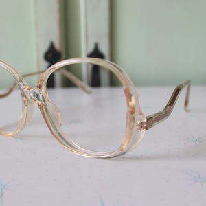 Peut inclure: Une paire de lunettes en plastique transparent avec une monture en métal doré. Les lunettes ont une forme de lentille ronde et surdimensionnée.