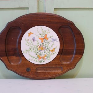 Puede incluir: Una bandeja de madera con una incrustación de baldosas de cerámica decorativa. La baldosa presenta un diseño de mariposas en tonos naranja, amarillo y morado, rodeadas de follaje verde. La bandeja tiene dos asas curvas.