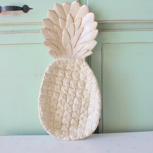 May include: A white ceramic pineapple-shaped serving dish with gold accents. The dish has a detailed design and is perfect for serving snacks or fruit.