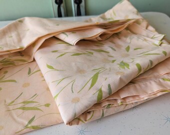 Peut inclure: Parure de lit pêche avec un motif de marguerites et de feuilles vertes. Les draps sont pliés, mettant en valeur le motif floral. Le tissu semble doux et léger, adapté à la literie.