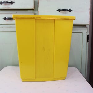 Könnte beinhalten: Eine leuchtend gelbe Kunststoff-Aufbewahrungsbox mit rechteckiger Form. Die Box hat einen Deckel und vertikale Rillen auf der Vorderseite. Die Box steht auf einer weißen Oberfläche, mit einem hellgrünen Schrank im Hintergrund.