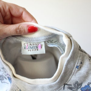 May include: A gray sweatshirt with a floral pattern in blue and black. The neckline is white with a gray trim. The tag reads "CLASSIC ELEMENTS PETITE".