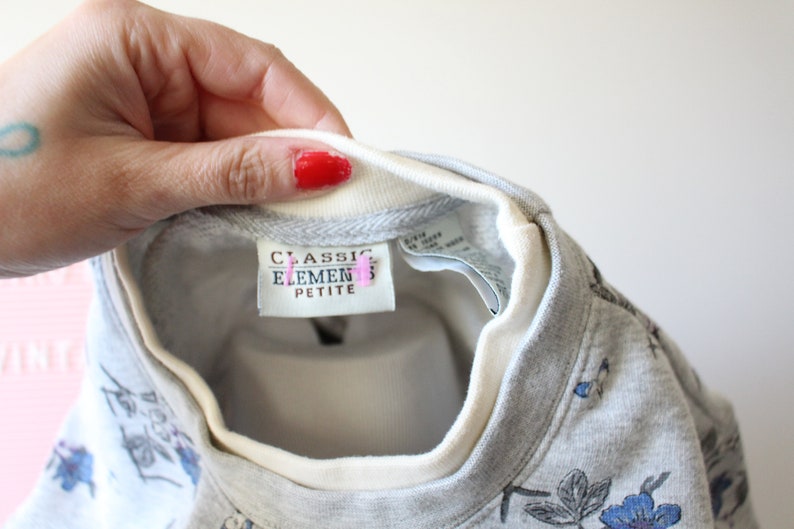 May include: A gray sweatshirt with a floral pattern in blue and black. The neckline is white with a gray trim. The tag reads "CLASSIC ELEMENTS PETITE".