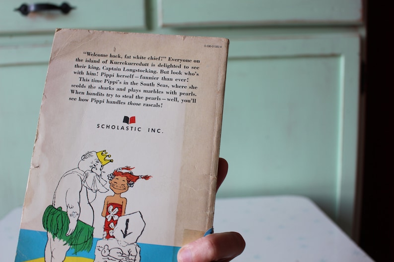 Peut inclure: La couverture arri&egrave;re d'un livre pour enfants illustrant un roi et une fille avec une couronne en dessin anim&eacute;. Le texte sur la couverture dit "Bienvenue, gros chef blanc !" Tout le monde sur l'&icirc;le de Kurrekurredutt est ravi de voir leur roi, le Capitaine Longstocking. Mais regardez qui est avec lui ! Pippi elle-m&ecirc;me - plus dr&ocirc;le que jamais ! Cette fois, Pippi est dans les mers du Sud, o&ugrave; elle gronde les requins et joue aux billes avec des perles. Quand des bandits essaient de voler les perles - eh bien, vous verrez comment Pippi s'occupe de ces voyous !" Le livre est publi&eacute; par Scholastic Inc.