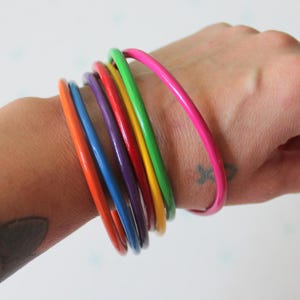 May include: A set of six thin, smooth, circular bangle bracelets in a vibrant rainbow color palette. The stack includes individual bangles in bright orange, blue, purple, red, yellow, green, and a larger pink bangle worn separately.