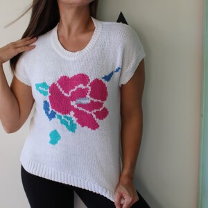 May include: White knitted sweater with a pink, blue, and green floral design. The sweater has short sleeves and a round neckline.