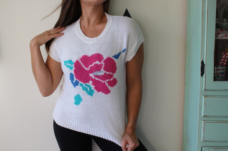 May include: White knitted sweater with a pink, blue, and green floral design. The sweater has short sleeves and a round neckline.