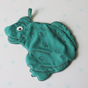 May include: A teal frog-shaped potholder with a loop for hanging. The potholder features a smiling frog face with a white eye and black stitching details. The frog has dark spots and a silver outline.