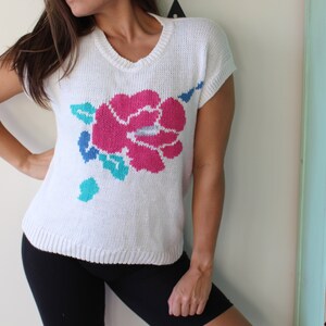 May include: White knitted sweater with a large pink flower design and green leaves. The sweater has short sleeves and a v-neck.