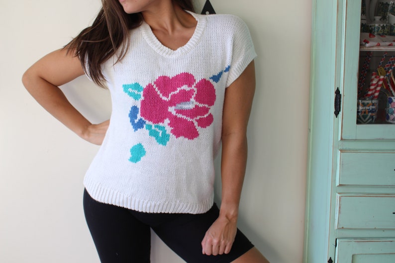 May include: White knitted sweater with a large pink flower design and green leaves. The sweater has short sleeves and a v-neck.