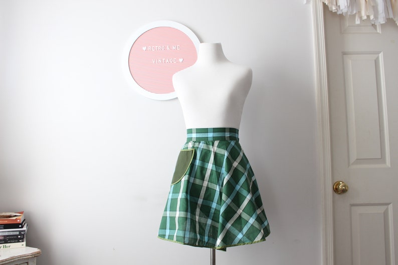 Pode incluir: Uma saia xadrez verde e azul com um bolso verde. A saia est&aacute; em um manequim branco e tem uma bainha amarela.