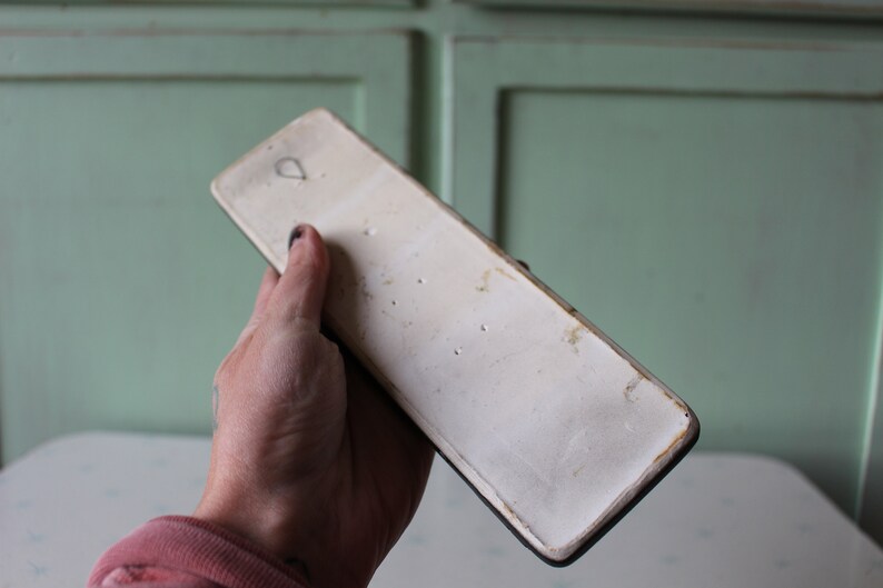 以下が含まれることがあります： 左上の角に小さな穴が開いた、白い長方形のセラミックタイルの裏側。タイルはわずかにざらざらした表面をしていて、濃い茶色の縁取りで囲まれています。