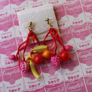 May include: A pair of dangle earrings with red string and colorful fruit charms, including a banana, cherries, and strawberries. The earrings are displayed on a white card with a pink and white background featuring Campbell's Soup cans.