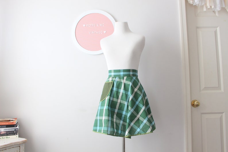 Pode incluir: Um avental xadrez verde e azul com um bolso verde. O avental est&aacute; em um manequim branco. O texto "Retro & Me Vintage" est&aacute; em uma placa rosa atr&aacute;s do manequim.