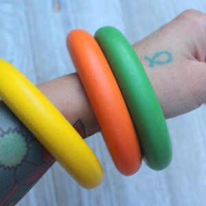 May include: Three smooth, solid-colored bangle bracelets in bright yellow, vibrant orange, and grass green. These circular accessories feature a rounded, thick profile and a matte finish, worn together as a set on a forearm.