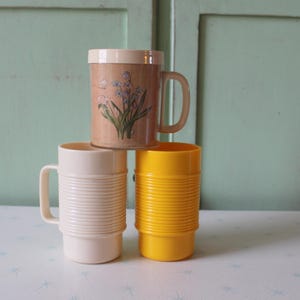 Könnte beinhalten: Drei Vintage-Tassen aus Kunststoff in Creme, Gelb und Hellbraun. Die hellbraune Tasse hat ein Blumenmuster. Jede Tasse hat einen Griff und ein geripptes Design um den Körper. Die Tassen sind übereinander gestapelt.