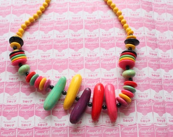 Könnte beinhalten: Eine bunte Halskette aus Holzperlen in verschiedenen Formen und Größen. Die Perlen sind in den Farben Gelb, Grün, Rot, Pink und Schwarz gehalten. Die Halskette liegt auf einem rosa-weißen Hintergrund mit einem sich wiederholenden Muster von Campbell's Soup-Etiketten.