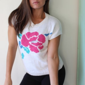 May include: White knitted sweater with a pink, blue, and green floral design. The sweater has short sleeves and a round neckline.
