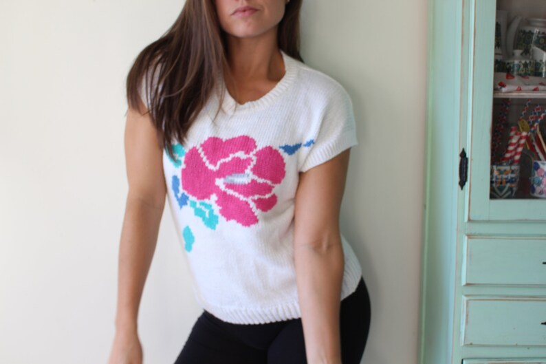 May include: White knitted sweater with a pink, blue, and green floral design. The sweater has short sleeves and a round neckline.