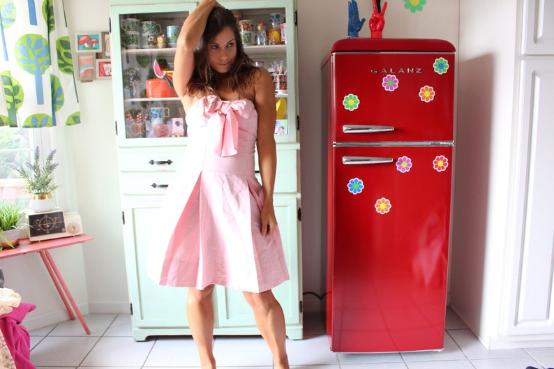 May include: A woman in a pink strapless dress with a bow at the neckline stands in a kitchen with a red refrigerator with flower stickers. The refrigerator has the word "GALANZ" on it.