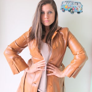 May include: A woman wearing a brown leather jacket with a belt. The jacket has a button closure and long sleeves. The woman is standing in front of a white wall.