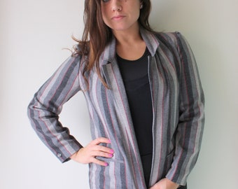 May include: Gray blazer with vertical stripes in red and white. The blazer has a notched collar, a single-button closure, and two front pockets. The sleeves are long, and the blazer is worn over a black top and black pants.