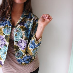 Puede incluir: Una chaqueta con estampado floral sobre fondo blanco con flores azules, moradas y verdes. La chaqueta tiene cuello y mangas largas.