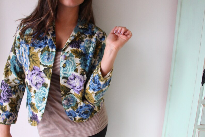 Puede incluir: Una chaqueta con estampado floral sobre fondo blanco con flores azules, moradas y verdes. La chaqueta tiene cuello y mangas largas.