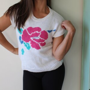 May include: White knitted sweater with a pink, blue, and turquoise flower design.