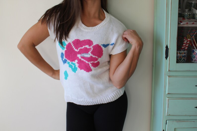 May include: White knitted sweater with a pink, blue, and turquoise flower design.