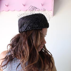 Puede incluir: Un sombrero negro acolchado con un velo delicado. El sombrero est&aacute; hecho de un material brillante. La mujer de la imagen tiene el pelo largo y ondulado de color marr&oacute;n y lleva una camiseta a rayas. Una estanter&iacute;a rosa es visible en el fondo.