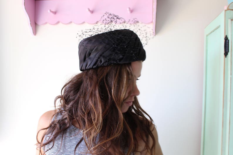 Puede incluir: Un sombrero negro acolchado con un velo delicado. El sombrero est&aacute; hecho de un material brillante. La mujer de la imagen tiene el pelo largo y ondulado de color marr&oacute;n y lleva una camiseta a rayas. Una estanter&iacute;a rosa es visible en el fondo.