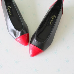 Puede incluir: Un par de zapatos planos de cuero negro con punteras de charol rojo. Los zapatos tienen una punta puntiaguda y un diseño sencillo. El nombre de la marca "Bijou" está impreso en la plantilla.