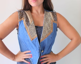 May include: A blue denim vest with tan and leopard print accents. The vest features a V-neck design with tan trim and gold-colored rivets. The shoulders have leopard print fabric, and the vest has a button closure.