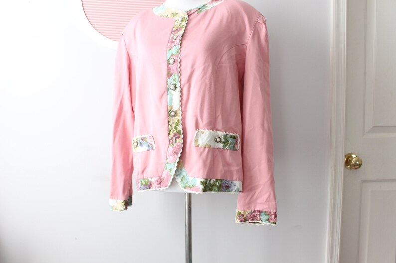 May include: A pink blazer with floral trim and a scalloped edge. The blazer has a button closure and two pockets.