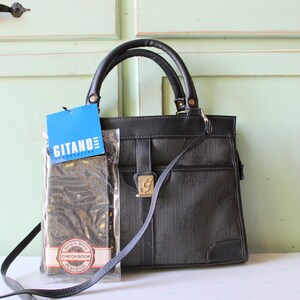 May include: Black handbag with a top handle and a detachable shoulder strap. The bag has a flap closure with a gold-colored clasp. A blue tag with the text "GITANO ACCESSORIES" is attached. A wallet is in a clear plastic sleeve.