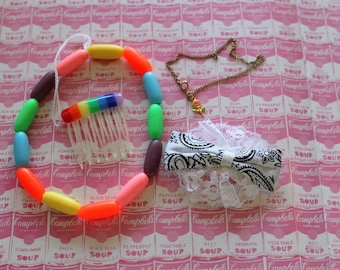 Könnte beinhalten: Eine bunte Perlenkette mit einem Regenbogenkamm und einer goldenen Kette mit einem Blumenanhänger. Die Kette liegt auf einem rosa und weißen Hintergrund mit einem Muster aus Campbell's Suppendosen.