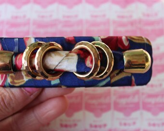May include: A blue and red patterned fabric hair clip with gold metal accents. The clip has a knot design with three gold rings.
