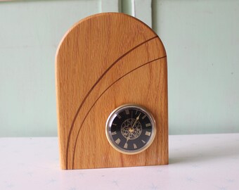 May include: A wooden desk clock with a light brown finish and a curved top. The clock face is black with gold-colored hands and Roman numerals. Decorative curved lines are etched into the wood.