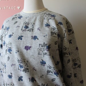 May include: A gray sweatshirt with a floral pattern. The design includes blue and white flowers, purple leaves, and gray leaves. The sweatshirt has a crew neck and long sleeves. The word "VINTAGE" is visible in the background.