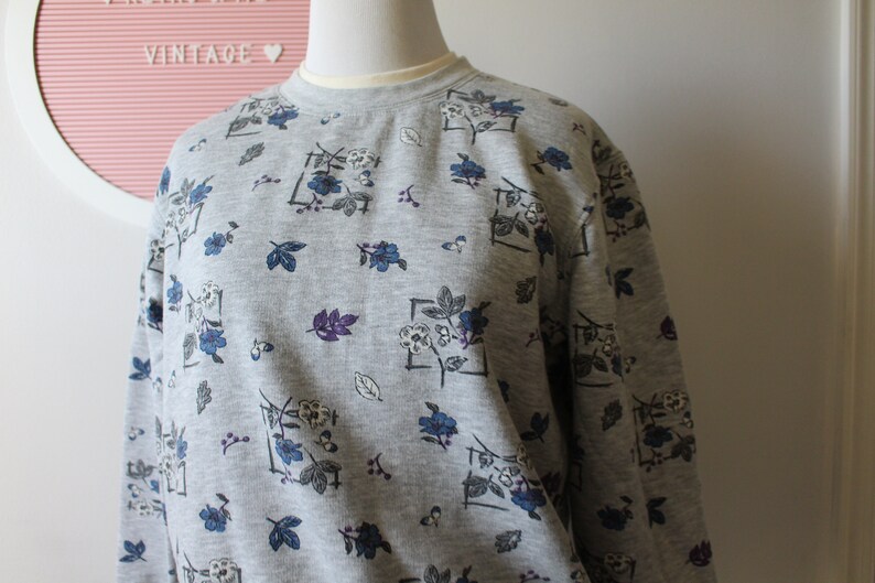 May include: A gray sweatshirt with a floral pattern. The design includes blue and white flowers, purple leaves, and gray leaves. The sweatshirt has a crew neck and long sleeves. The word "VINTAGE" is visible in the background.