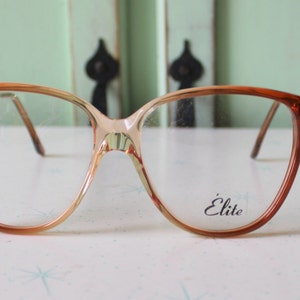 Puede incluir: Gafas de pasta de color marrón y beige con forma de ojo de gato, con lentes transparentes y la palabra "Elite" impresa en la lente derecha.