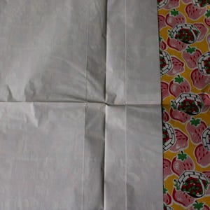 May include: A white sheet of paper folded in half with a yellow and pink patterned fabric on the right side. The fabric has a repeating pattern of strawberries in baskets.