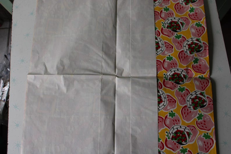 May include: A white sheet of paper folded in half with a yellow and pink patterned fabric on the right side. The fabric has a repeating pattern of strawberries in baskets.