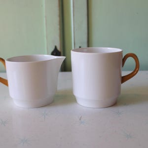 Könnte beinhalten: Ein weißes Keramik-Sahnekännchen und Tassen-Set mit braunen Griffen. Das Sahnekännchen hat einen Ausguss, und beide Teile haben ein schlichtes, modernes Design. Das Set wird auf einer weißen Oberfläche mit einem dezenten Muster präsentiert.